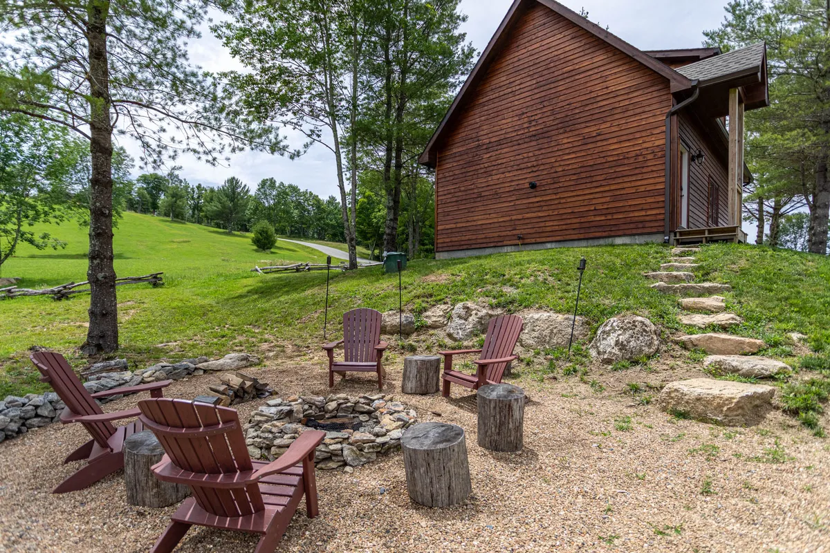 This inviting outdoor scene features a stone fire pit surrounded by Adirondack chairs, set beside a wooden cabin and bordered by a stone pathway. The area is clearly designed for campfire gatherings, relaxation, and enjoying the mountain air.