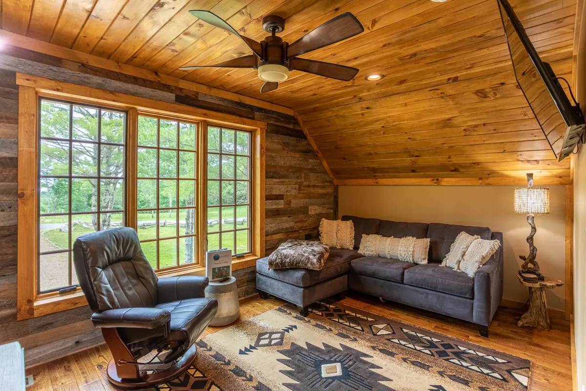 A sunlit living area featuring expansive windows that flood the room with natural light. The plush sectional sofa, decorative throw pillows, and unique area rug create a cozy yet elegant atmosphere. A reclining chair and ambient lamp enhance the relaxing vibe of the space.