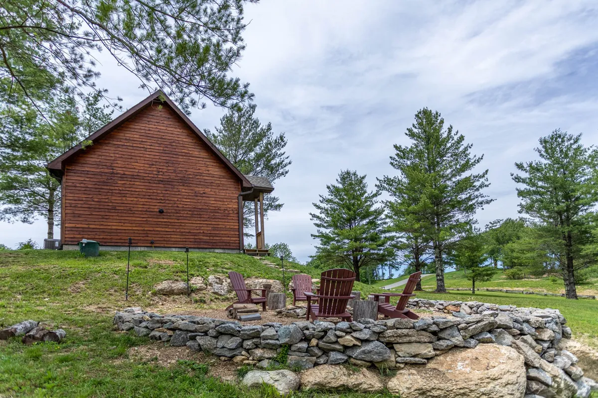 A peaceful outdoor setup showing a wooden cabin, a round stone fire pit, and several Adirondack chairs nestled among tall trees. The scene radiates tranquility and invites guests to enjoy both daytime nature and starlit nights.