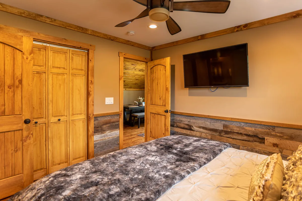 A calm and welcoming master bedroom featuring a plush bed with a textured blanket, wooden doors letting in natural sunlight, and a flat-screen TV mounted on the wall. The space is styled with soft, neutral tones and simple decorative touches that create a relaxing atmosphere.