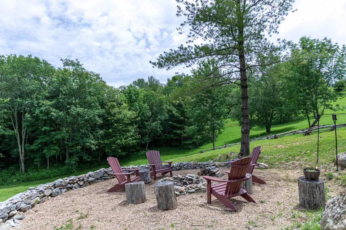 A cozy fire pit area framed by red Adirondack chairs and rustic stump seats, surrounded by lush green trees. The intimate outdoor setting invites guests to gather, unwind, and enjoy the sights and sounds of nature, whether for evening storytelling or quiet reflection.