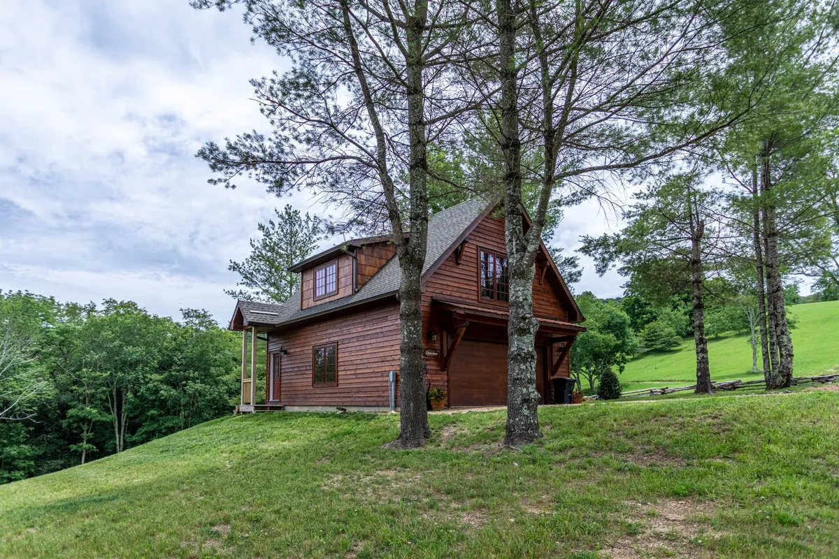 A peaceful view of a wooden cabin nestled among tall trees on a gentle grassy slope. The inviting porch faces out to the forest, suggesting a seamless connection with nature. The area exudes tranquility, perfect for outdoor relaxation, stargazing, or enjoying a quiet morning coffee.