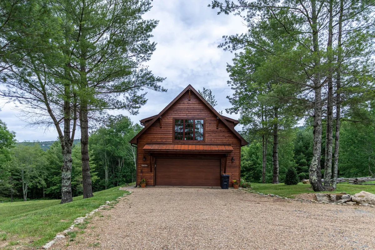 This exterior image features a wooden structure with expansive windows, a gravel driveway, and surrounding trees, creating a tranquil and inviting atmosphere. The view hints at the property's natural setting and the opportunities for outdoor activities and gatherings.