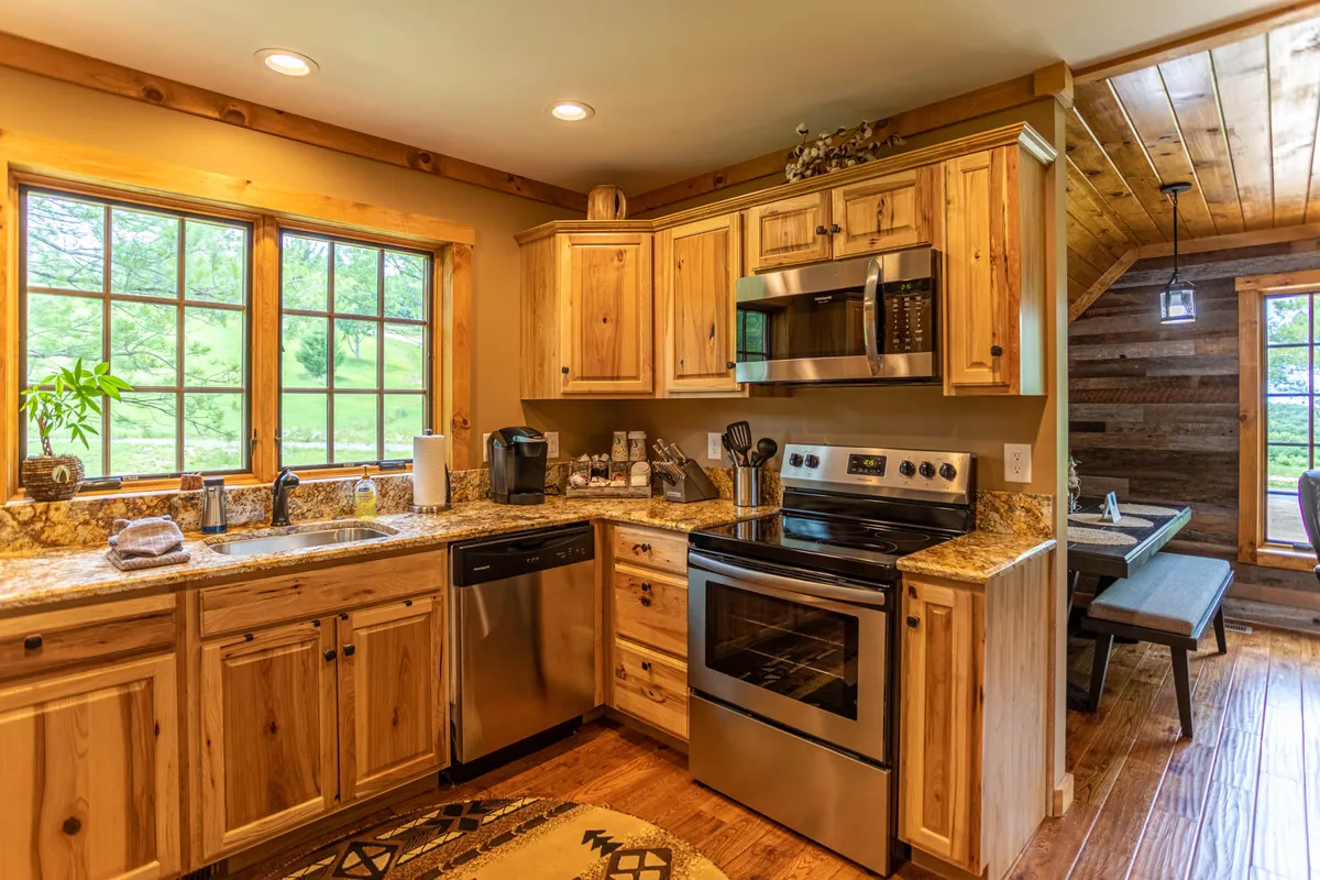 A warm and inviting kitchen area featuring wooden cabinetry, sleek granite countertops, stainless steel appliances, and large windows that let in natural light. The space is designed for both casual dining and culinary creativity, offering views of nature outside while providing all the amenities needed for meal preparation and entertaining.