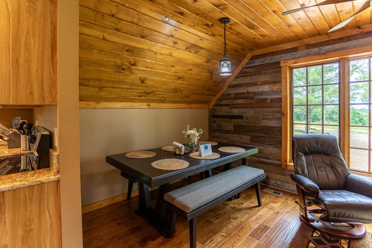 A cozy dining area anchored by a wooden dining table set for six, with a recliner chair nearby and a window offering a sunrise view. The space exudes warmth and is ideal for shared meals, casual gatherings, or simply enjoying the landscape beyond the glass.